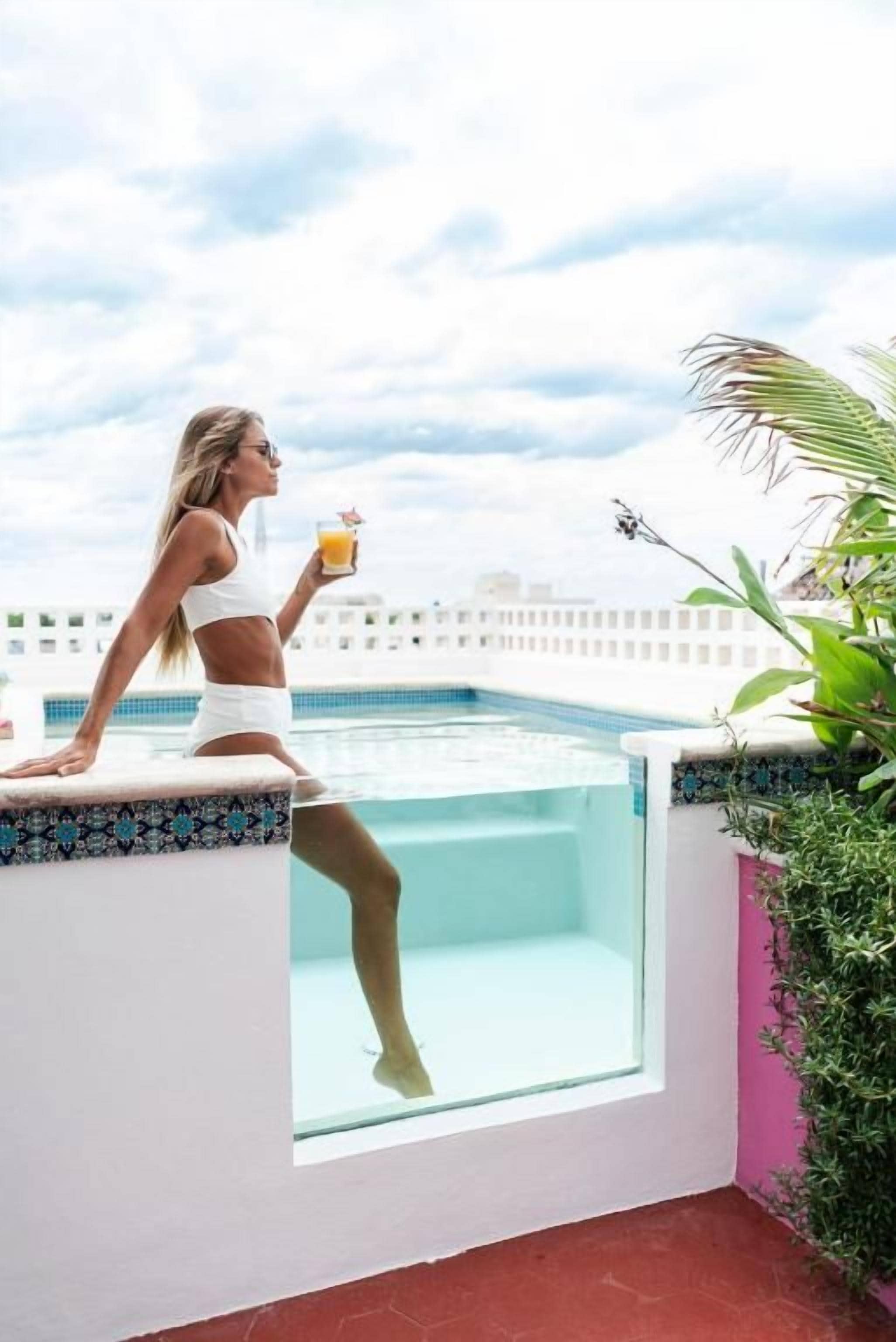 Una persona sentada junto a una piscina con una bebida, rodeada de follaje y un cielo despejado.