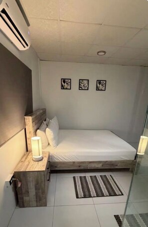Ein Einzelbett mit hölzernem Kopfteil, ein Nachttisch mit Lampe sowie eine Wand-Klimaanlage.