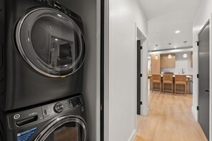 Een wasserette met twee voorlaadwasmachines en drogers, een gang naar een keuken met houten kasten en hanglampen.