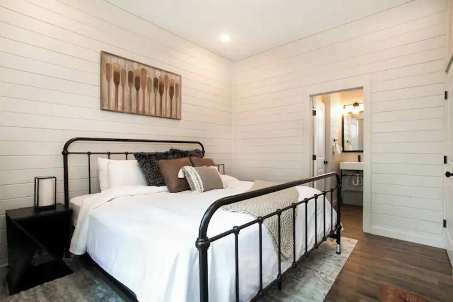 Un dormitorio con estructura de cama metálica, ropa de cama blanca, una mesita de noche, una obra de arte colgada en la pared y vistas al baño.