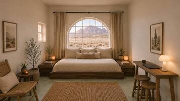 A bedroom with a large bed, a desk, two chairs, a window with a desert view, and a small table with a lamp.