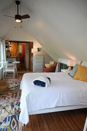 Ein Schlafzimmer mit einem großen Bett, einem Schreibtisch und einem Sessel. Es gibt einen Deckenventilator, eine Lampe und ein Fenster.