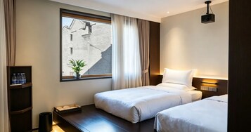 Habitación de hotel con dos camas, un ventanal grande con cortinas, una pequeña planta, un estante con botellas de agua y un proyector en el techo.