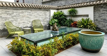 Una mesa de vidrio con sillas y una planta grande en un patio con paredes de ladrillo y plantas en macetas.