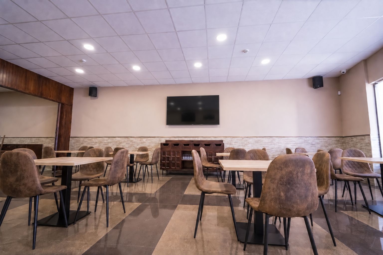 Un salón de conferencias con varias mesas y sillas, un televisor de pantalla plana montado en la pared y un mueble de madera con estantes.