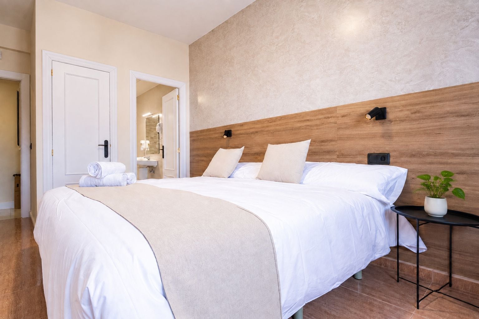 Quarto de hotel com uma cama grande, mesa de cabeceira com uma planta e banheiro visível através de uma porta aberta.