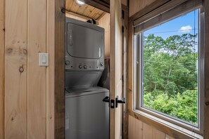 A compact, wooden interior with a built-in washer and dryer, a window with a view of greenery, and a door with a handle. - Lake View Cabin · Sleeps 16 · Hot Tub · Games (Sevierville)