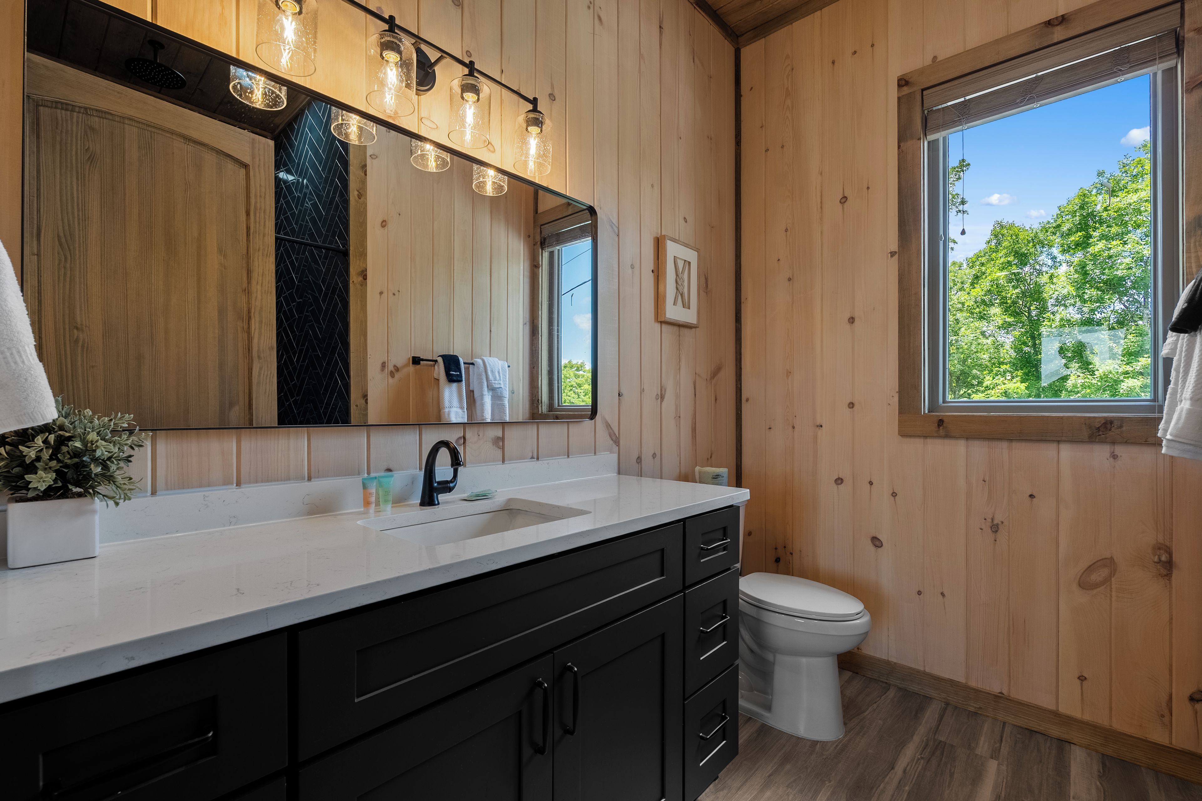 A bathroom with a wooden interior, a white sink, black cabinets, a toilet, and a window with a view of greenery.