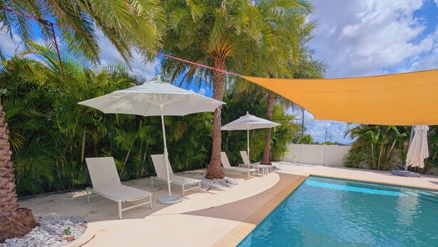 Ein Pool mit Liegestühlen und Sonnenschirmen, umgeben von Palmen und wolkenlosem Himmel.