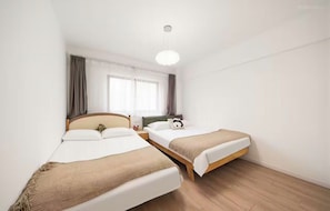 A hotel room with two beds, a window with curtains, a bedside table with a lamp, and a ceiling light.