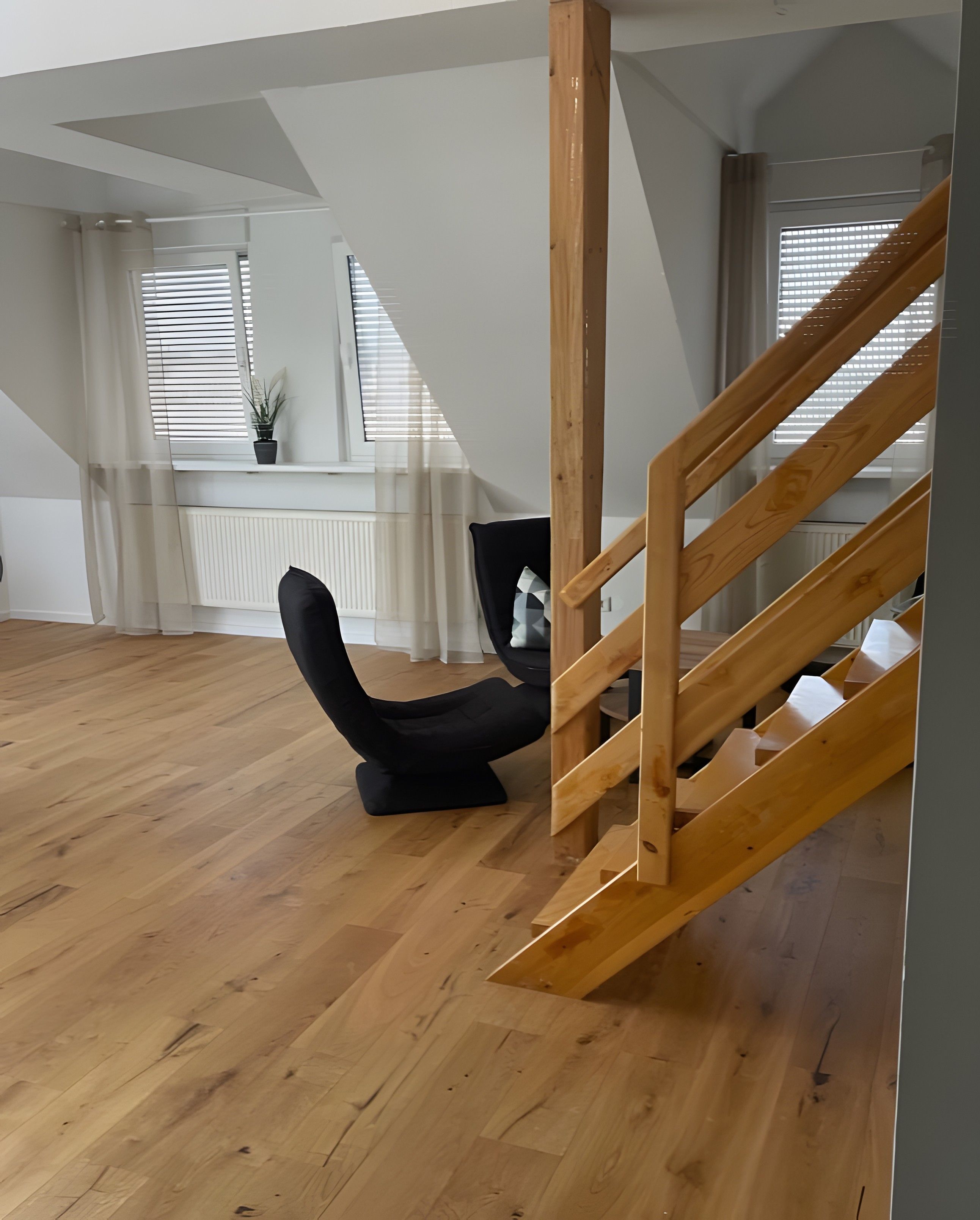 Quarto espaçoso com escada de madeira, uma cadeira preta e janelas amplas com persianas.