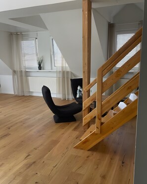 A spacious room with a wooden staircase, a black chair, and large windows with blinds.