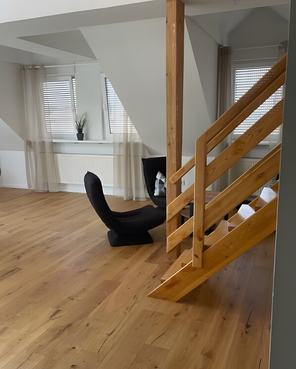 A spacious room with a wooden staircase, a black chair, and large windows with blinds.