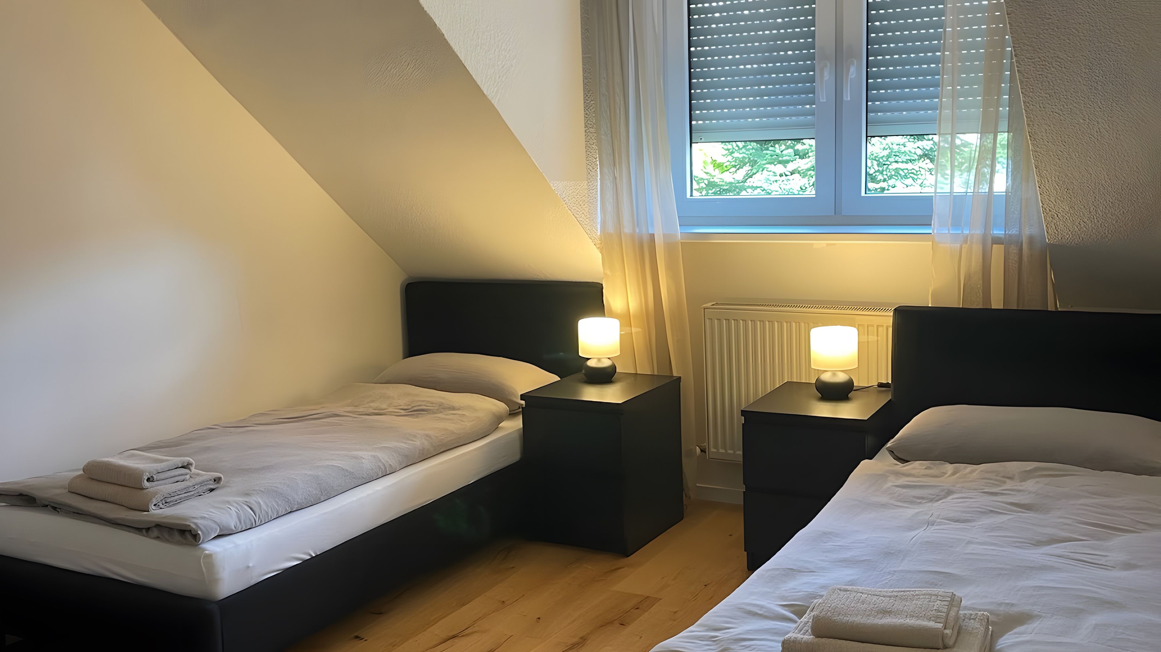 Ein Schlafzimmer mit zwei Betten, einem Fenster mit Jalousien, einem Heizkörper und Nachttischlampen.