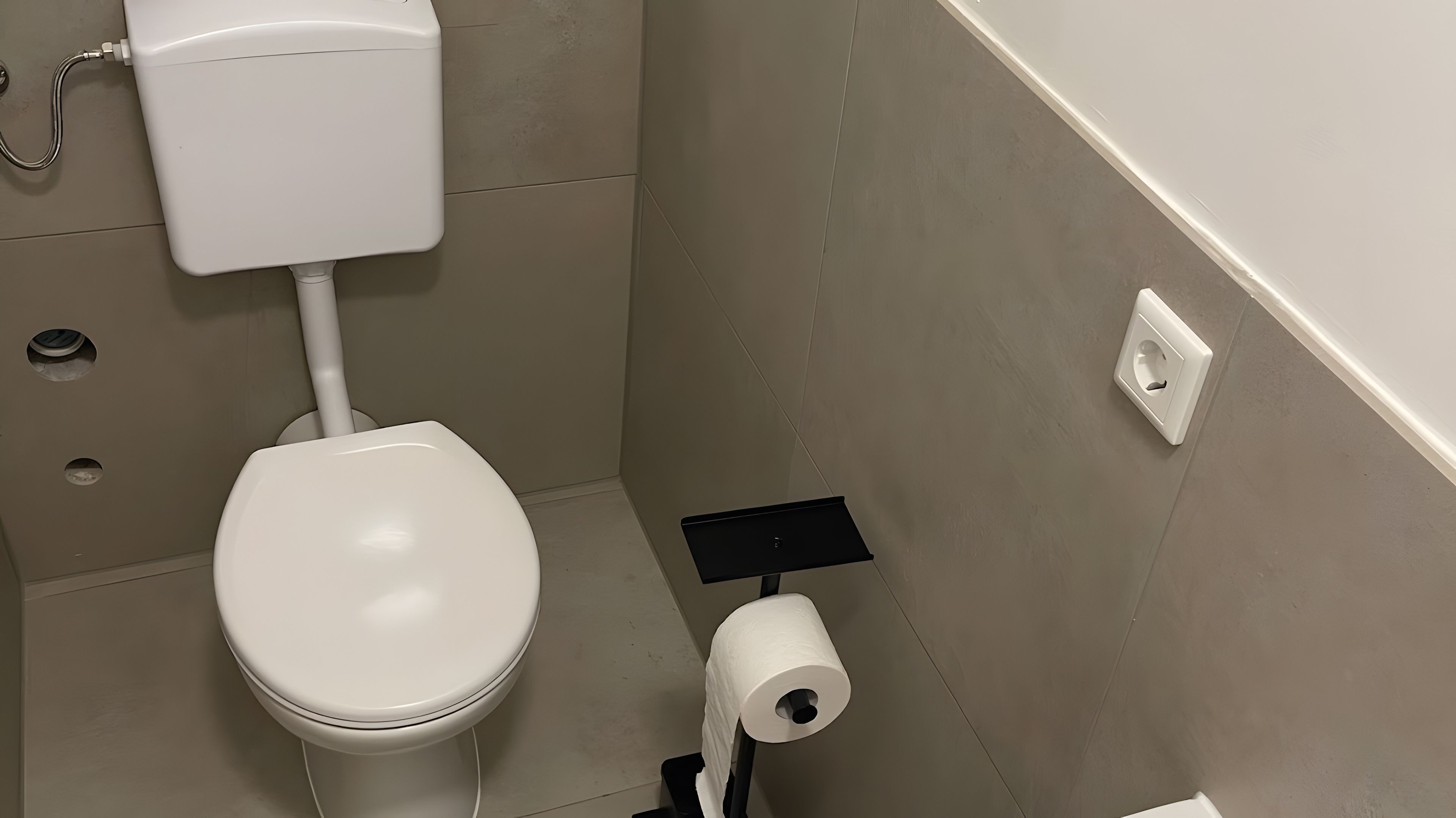 Eine Toilette mit weißem Tank und Deckel, ein Papierhandtuchhalter mit einer Rolle sowie eine an der Wand befestigte Steckdose.