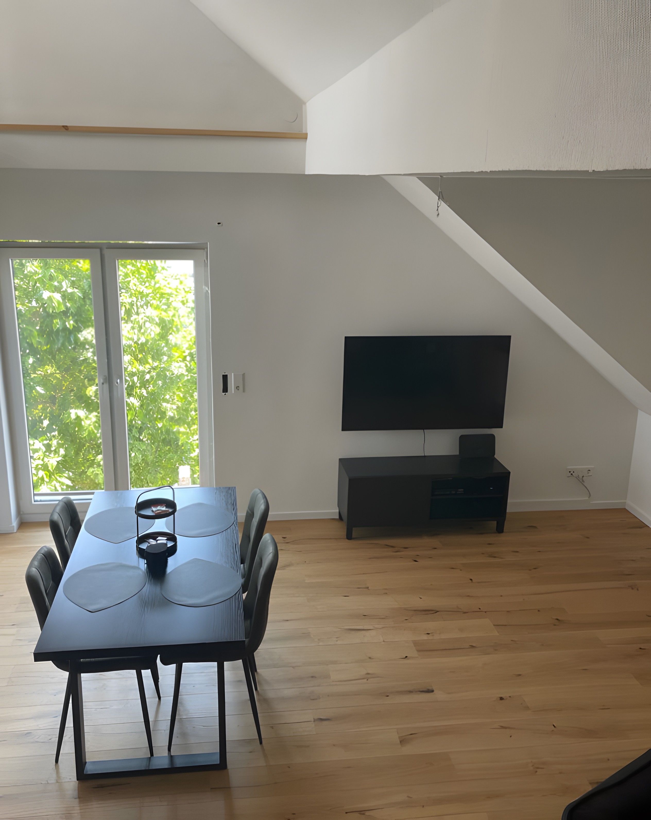 Un espace repas moderne avec une table et des chaises, un téléviseur à écran plat fixé au mur et une grande fenêtre donnant sur un paysage verdoyant.