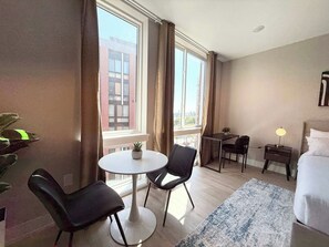 A hotel room with a bed, a round table with chairs, a small desk with a chair, a lamp, and a view of buildings through large windows. - Luxury Studio | 1 Bed | Northern Liberties (Philadelphia)