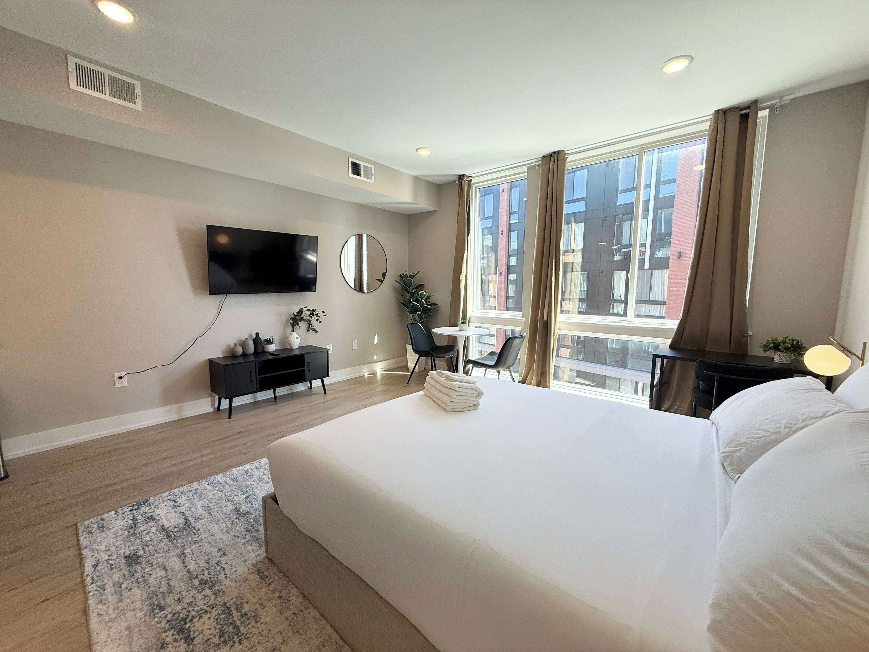 A hotel room with a large bed, a flat-screen TV mounted on the wall, a small desk with a chair, and a window with curtains.