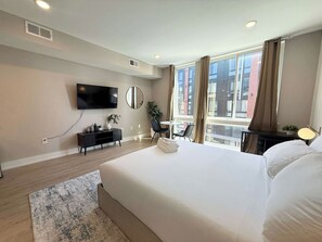 A hotel room with a large bed, a flat-screen TV mounted on the wall, a small desk with a chair, and a window with curtains. - Luxury Studio | 1 Bed | Northern Liberties (Philadelphia)