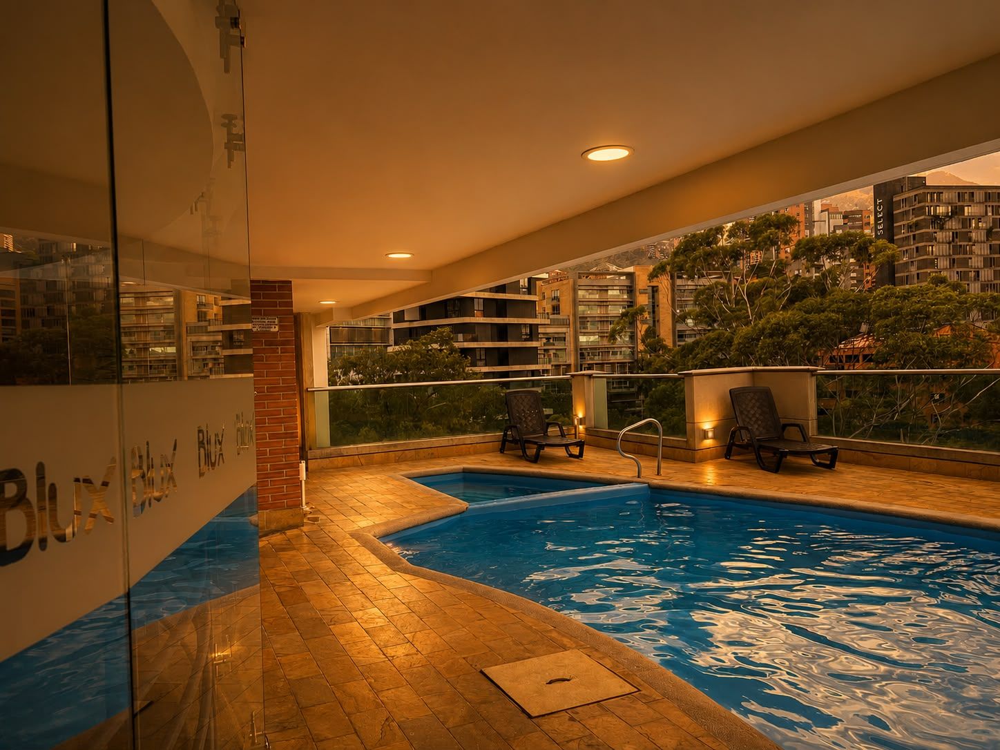 Piscina interna com parede de vidro, duas espreguiçadeiras e vista para edifícios ao redor.