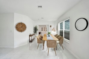 A modern dining area with a wooden table and wicker chairs, white walls, and a large round clock. - Coastal Bay Retreat | Pool | 8 Min to Beach (Fort Lauderdale)