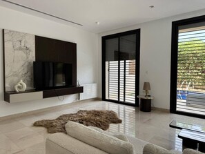 A modern living room with a large flat-screen TV mounted on the wall, a fur rug on the floor, and a view of the outdoors through large windows. - Malaga, duplex (Tunis)