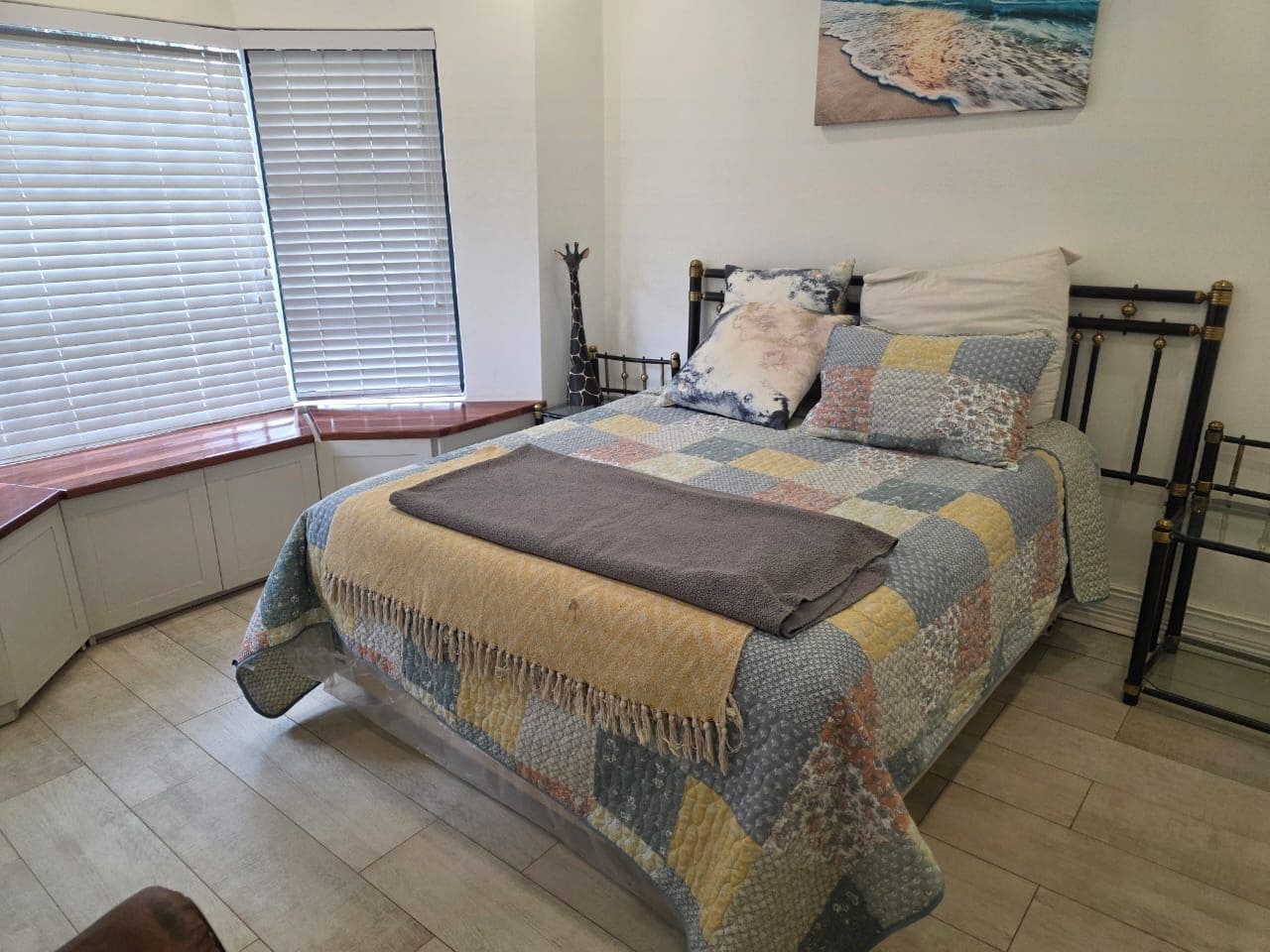 Un dormitorio con cama, ventana con persianas, un cuadro colgado en la pared y una mesita de noche.