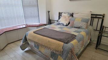 Un dormitorio con cama, ventana con persianas, un cuadro colgado en la pared y una mesita de noche.