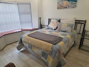 A bedroom with a bed, a window with blinds, a wall-mounted painting, and a bedside table. - Charles Olley  (Harare)
