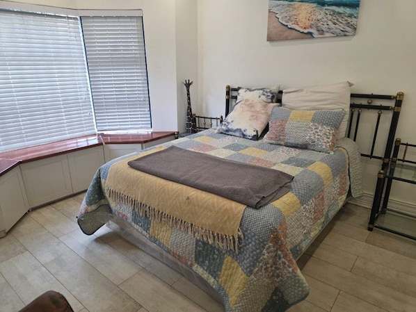 A bedroom with a bed, a window with blinds, a wall-mounted painting, and a bedside table.