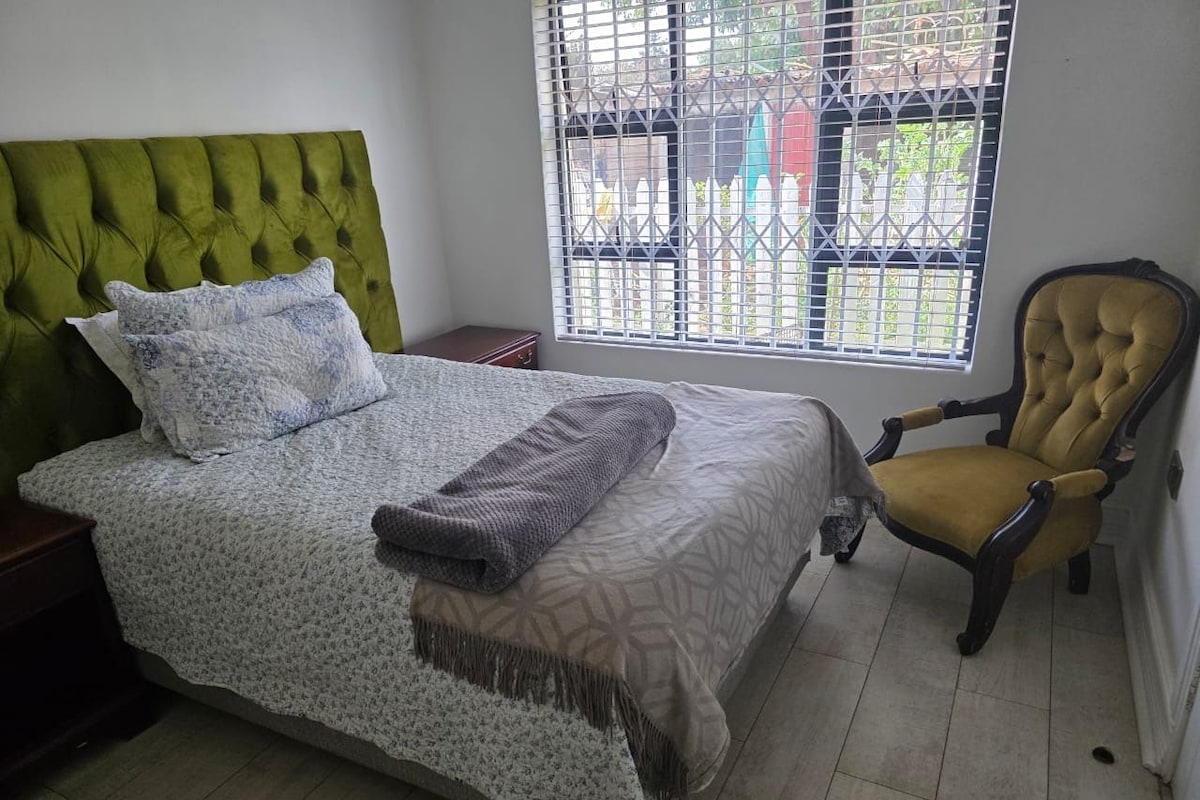 A bedroom with a bed, a chair, a window with a view, and a bedside table.