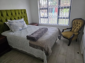 A bedroom with a bed, a chair, a window with a view, and a bedside table. - Charles Olley  (Harare)
