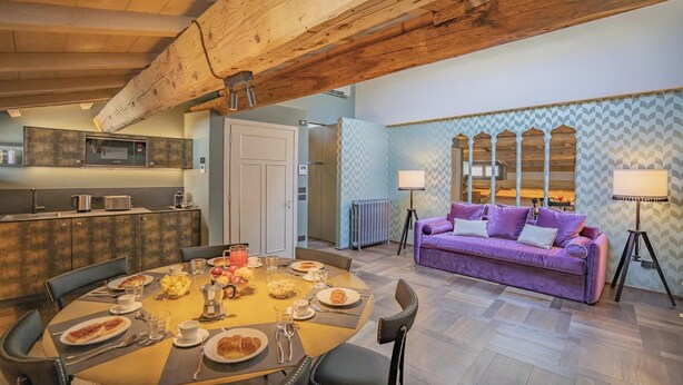 Un comedor con una mesa redonda dispuesta para el desayuno, un sofá morado y una cocina al fondo.