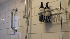 Eine Dusche mit Handbrause, flexiblem Duschschlauch und wandmontiertem Wasserhahn. Eine Seifenspender und eine Shampoo-Flasche befinden sich auf einem Metallregal.