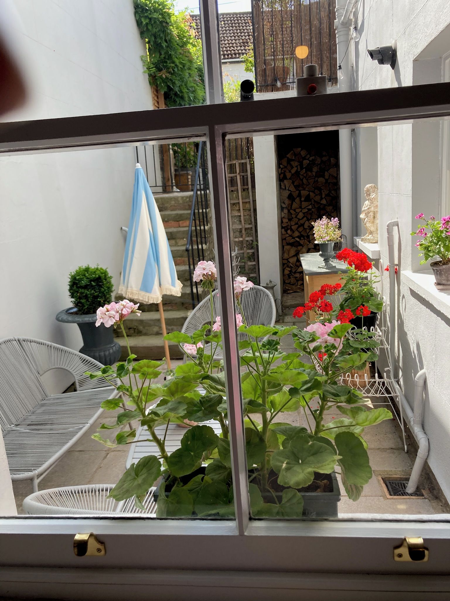 A view through a window of a balcony with potted plants, a chair, and a statue.