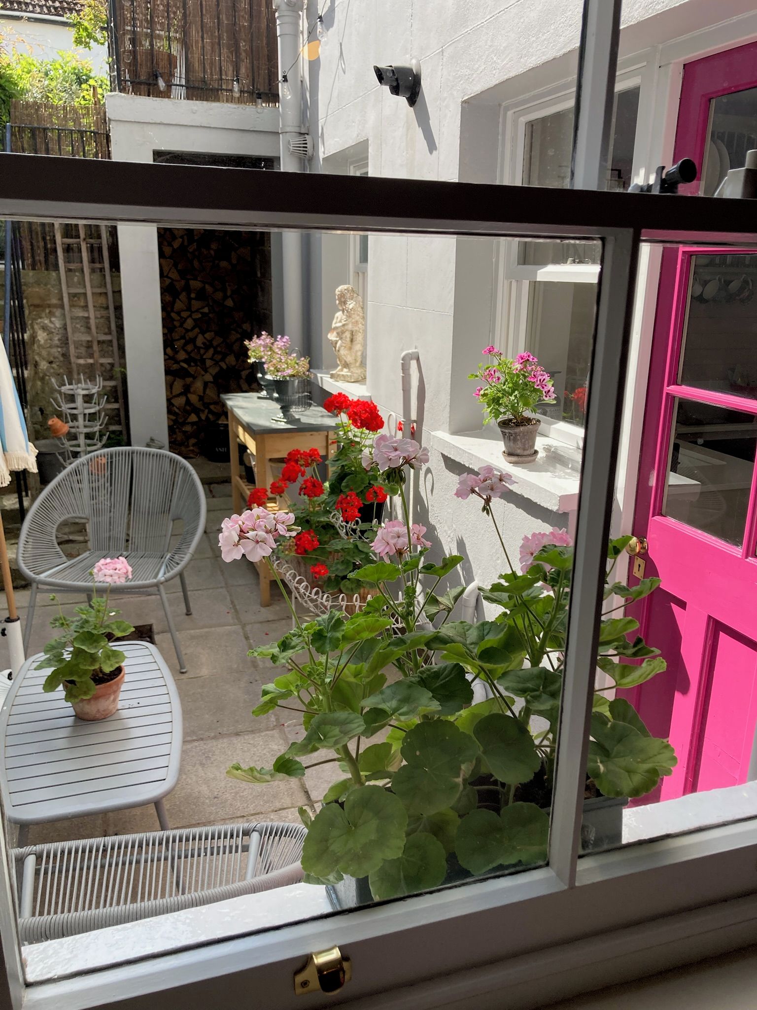 Uitzicht door een raam op een patio met een witte deur, een stoel, een tafel en potplanten.