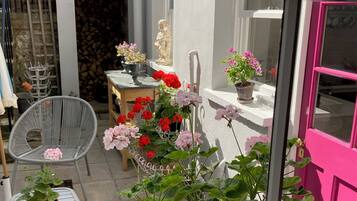 Uitzicht door een raam op een patio met een witte deur, een stoel, een tafel en potplanten.