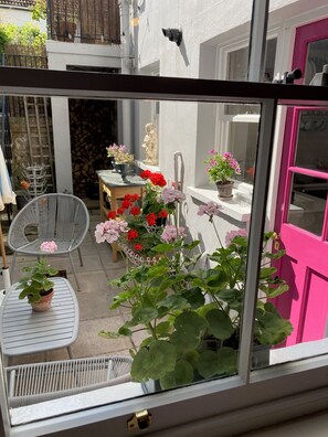Vista a través de una ventana que muestra un patio con una puerta blanca, una silla, una mesa y plantas en macetas.