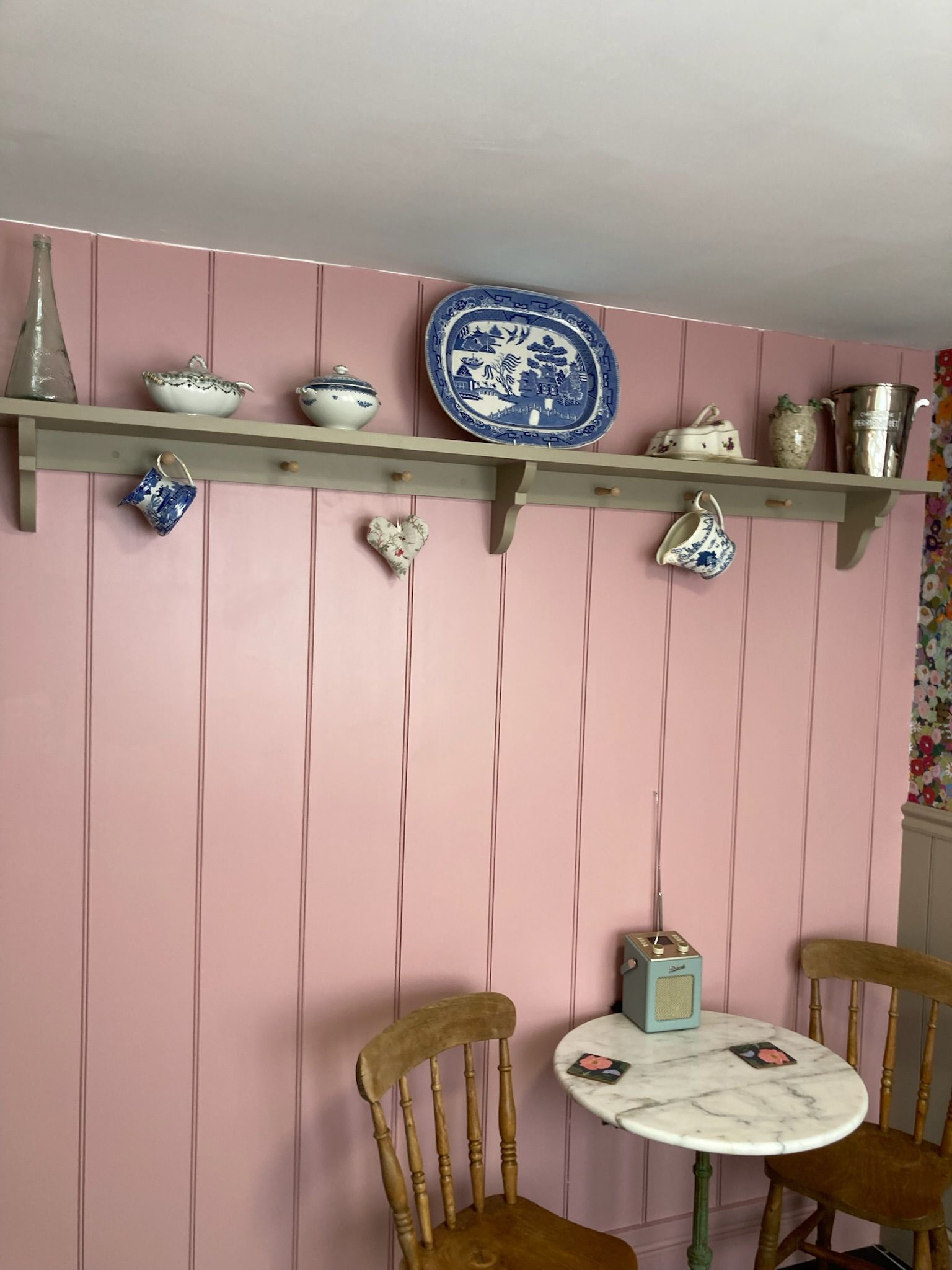 Een kamer met een roze wand, een houten plank met diverse keramieken objecten, een klein rond tafeltje met stoelen en een vintage radio.