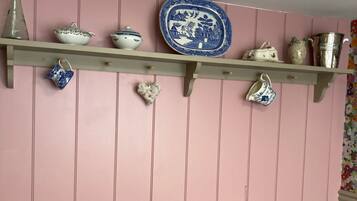 Een kamer met een roze wand, een houten plank met diverse keramieken objecten, een klein rond tafeltje met stoelen en een vintage radio.