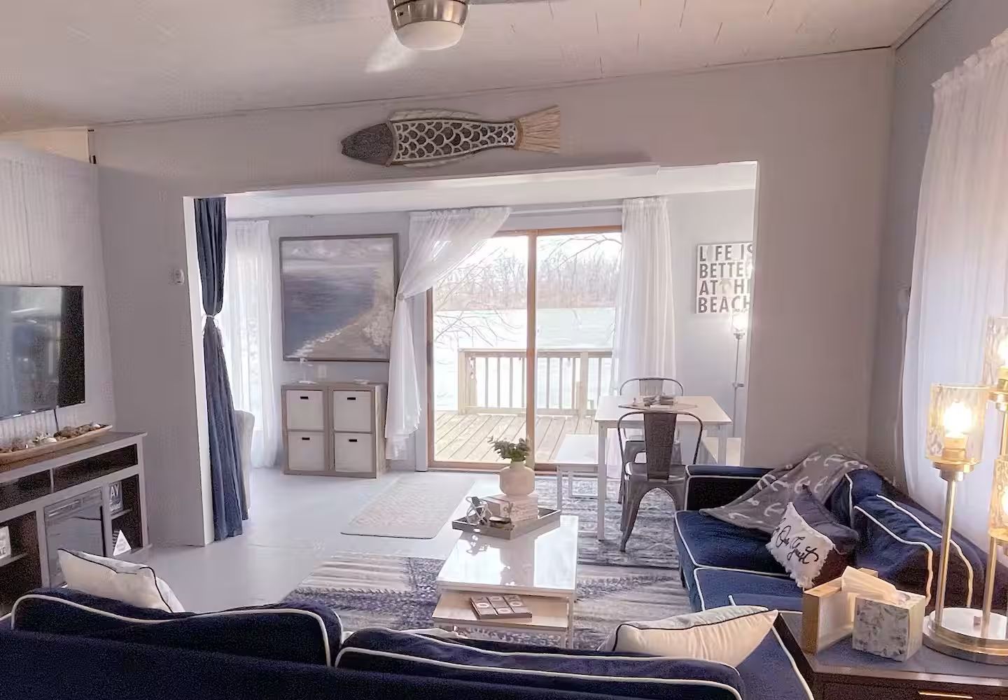 Una sala de estar con sofá, mesa de centro y televisor. Se ve un ventanal con cortinas y vistas al exterior.