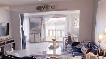 Una sala de estar con sofá, mesa de centro y televisor. Se ve un ventanal con cortinas y vistas al exterior.