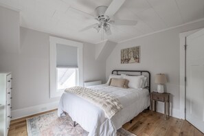 Ein Schlafzimmer mit einem Bett, einem Nachttisch, einem Deckenventilator, einem Fenster mit Jalousien und einer Heizung.