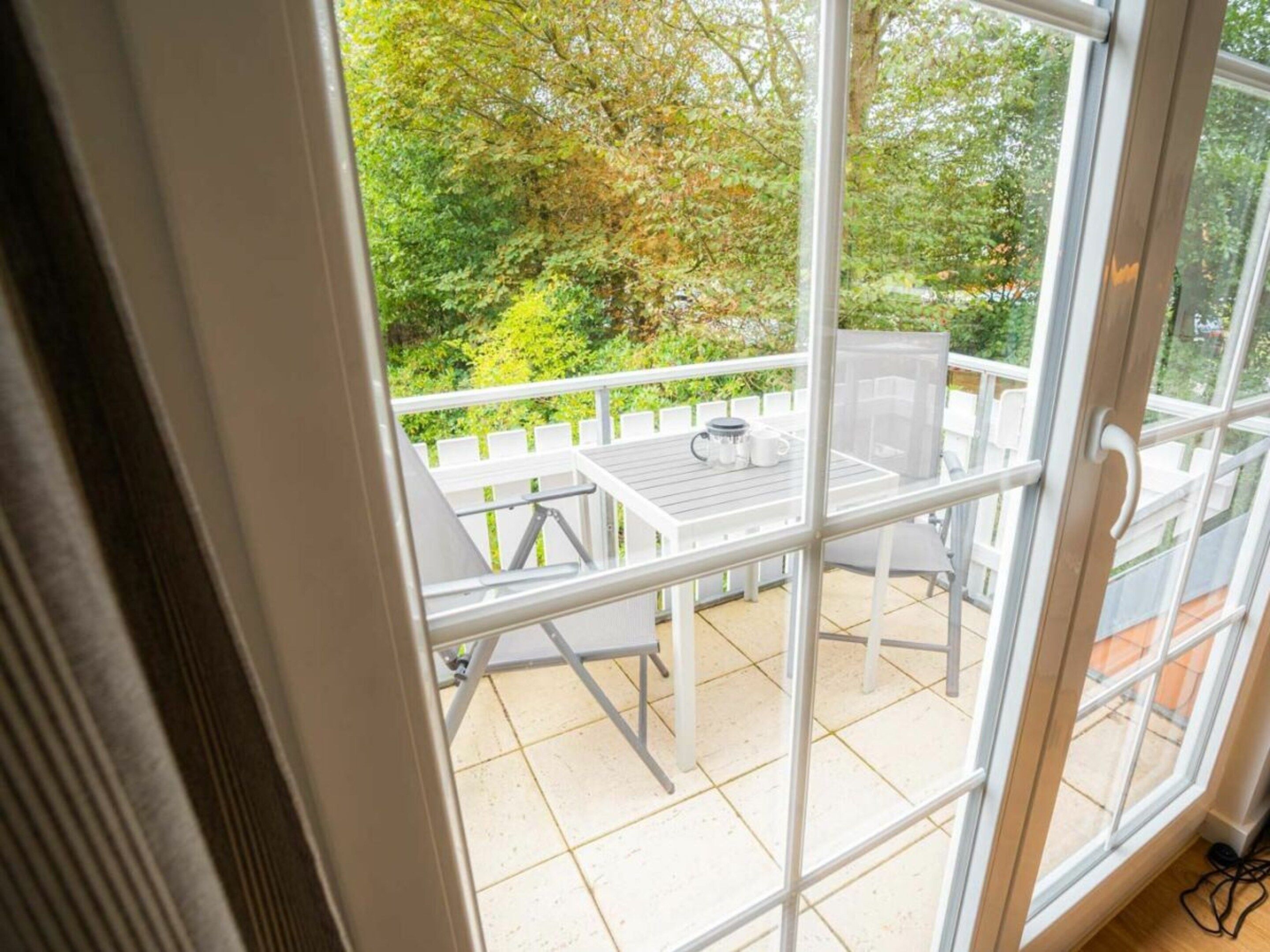 Vista através de uma porta de vidro para um pátio externo com mesa e cadeiras brancas, cerca branca e vegetação ao fundo.