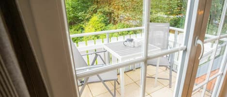 Vista através de uma porta de vidro para um pátio externo com mesa e cadeiras brancas, cerca branca e vegetação ao fundo.
