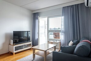Uma sala de estar moderna com uma televisão de tela plana em um suporte de madeira, uma mesa de centro clara, um sofá cinza e uma janela com cortinas cinzas.