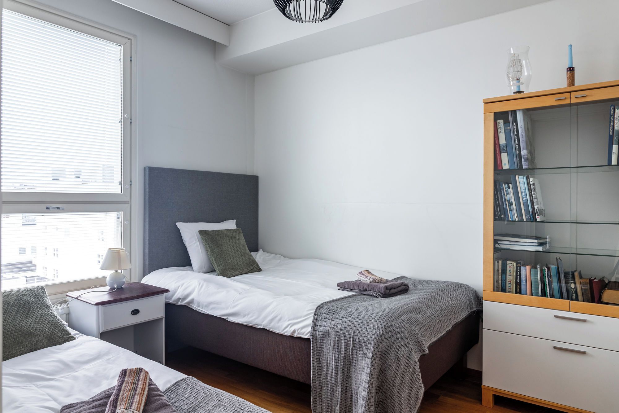 A bedroom with two beds, a bedside table with a lamp, a bookshelf, and a window with blinds.