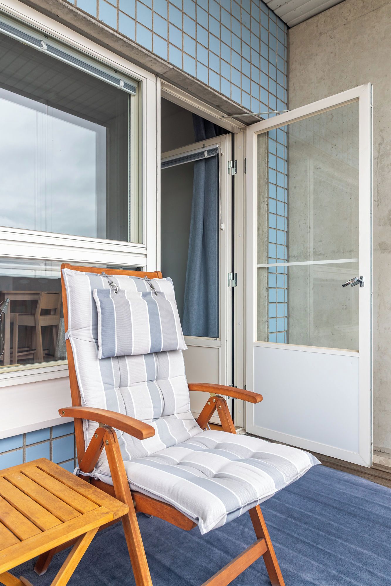 A wooden chair with striped cushions, a wooden table, and a view of a building with blue-tiled walls and a door.