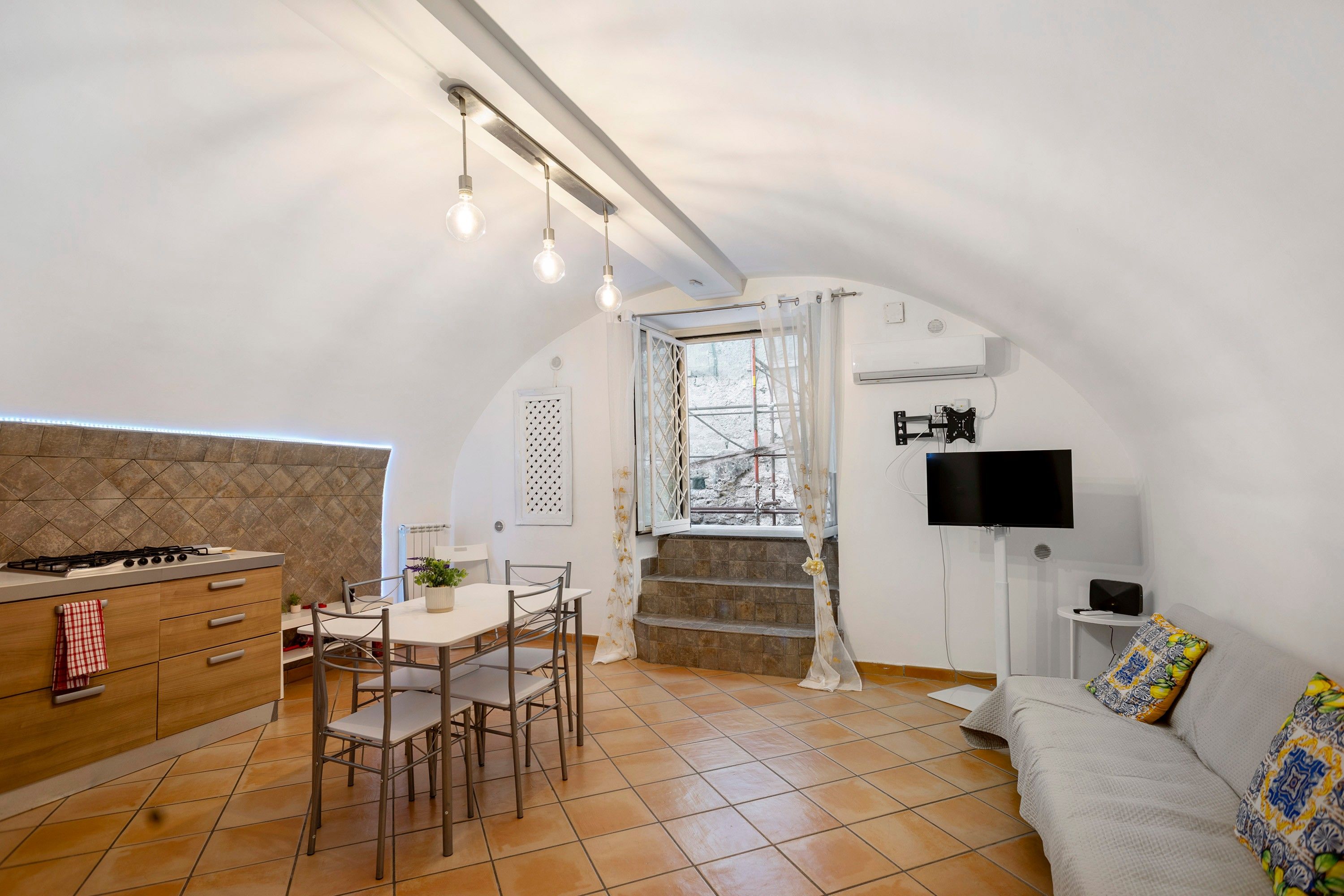 A compact kitchen with a gas stove, dining table, and chairs. There is a TV mounted on the wall and a sofa in the living area.