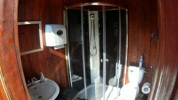 Baño de cabaña de madera con ducha acristalada, lavamanos, inodoro y dispensador de jabón.